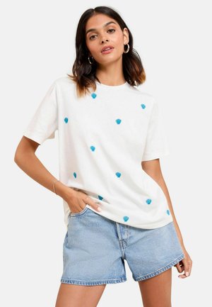 T-shirt en coton blanc avec broderie en coquillage bleu, manches courtes, col rond. Porté avec un short en denim léger, mettant en avant une coupe décontractée et un style casual.