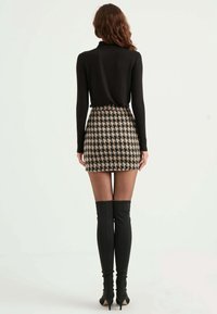 Frau steht mit dem Rücken zur Kamera und trägt ein schwarzes Langarmoberteil, einen Hahnentrittmuster-Minirock, transparente Strumpfhosen und schwarze kniehohe Stiefel mit Absatz.