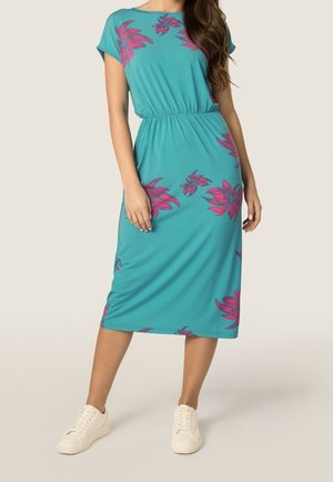 Vrouw die een turquoise midi-jurk draagt met roze bloemenprint en witte sneakers, staand met één voet iets naar voren.
