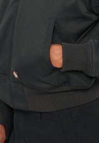 Melna bumbierjaka ar ribotu apakšmalu un piedurknēm, ar ārējo kabatu. Gluda auduma tekstūra ar mazu logotipa etiķeti sānos.
