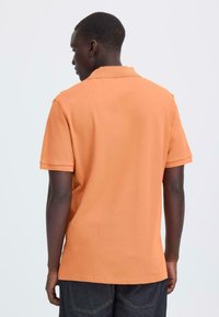 Polo à manches courtes orange en tissu coton, avec un col décontracté et un ourlet droit, associé à un jean foncé. Vue de dos.