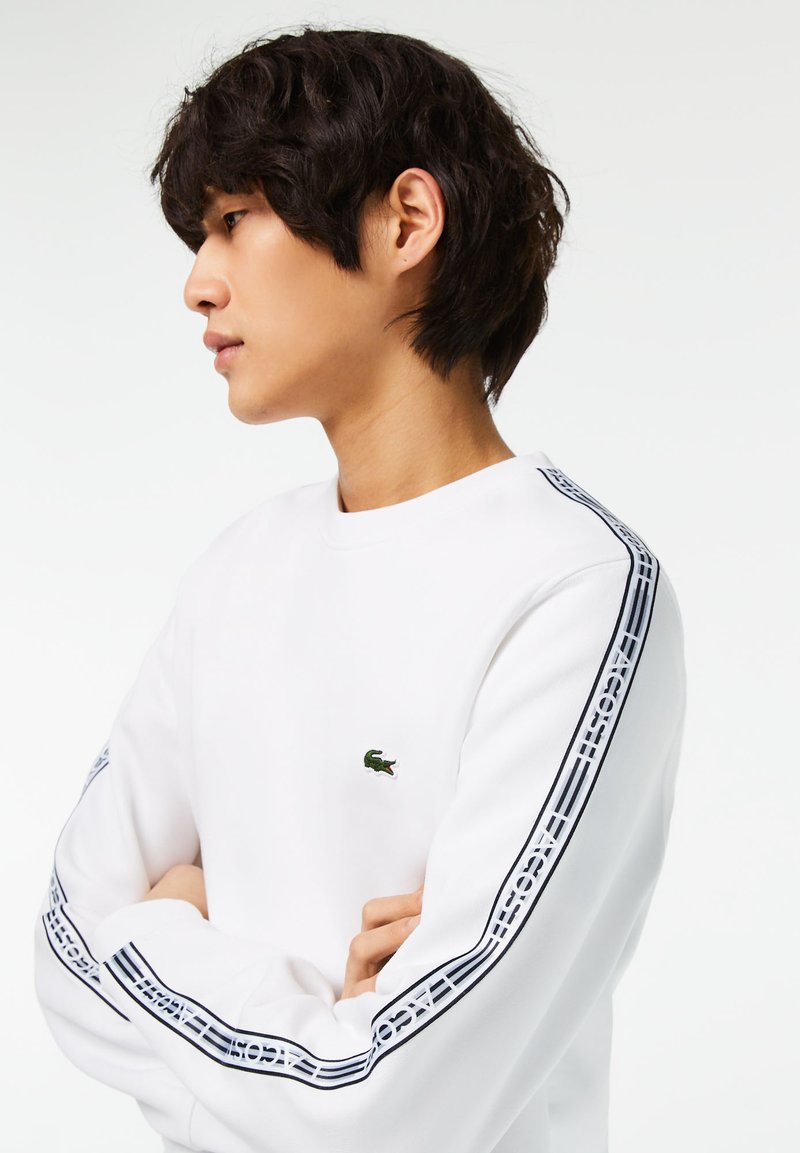 Sweatshirt blanc avec des accents de rayures noires le long des manches, comportant un petit logo vert sur la poitrine. Texture de tissu douce, design à col rond.