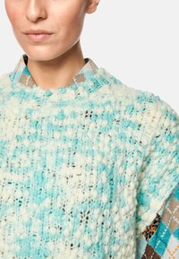 Light blue knitted pullover with textured bobbles, layered over a patterned shirt featuring geometric and animal prints.