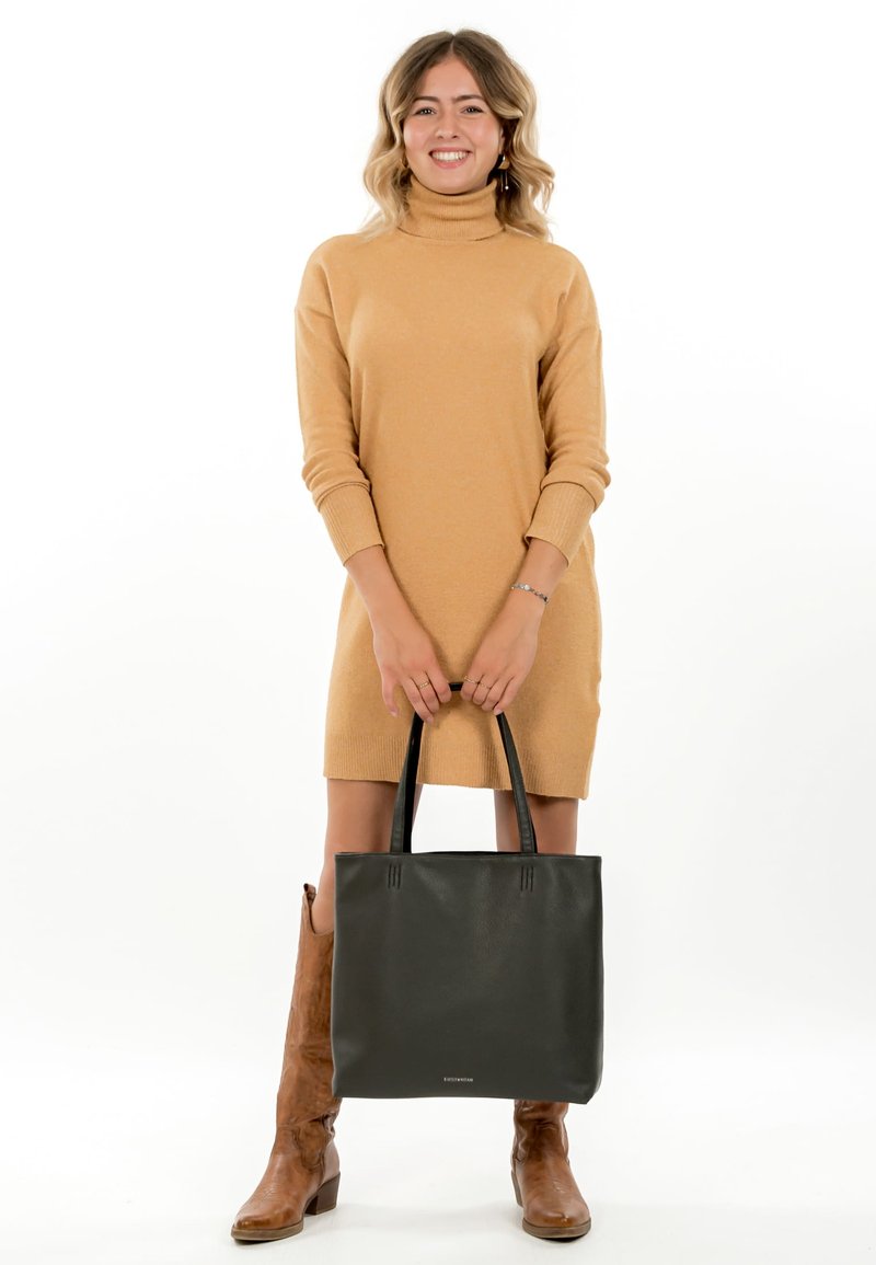 Femme souriante, portant une robe col roulé beige et des bottes marron, tenant un grand sac cabas en cuir noir à deux mains devant elle.