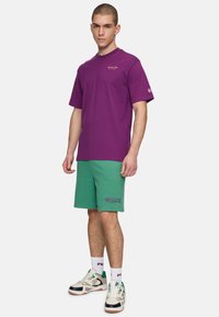Paarse katoenen t-shirt met korte mouwen en een ronde hals, gecombineerd met groene shorts met paarse logo-tekst. Sneakers met multikleurige accenten.