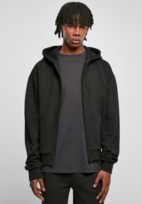 Sweatshirt zippé noir en tissu texturé avec capuche, comportant deux poches avant et des poignets côtelés, superposé à une chemise gris foncé.