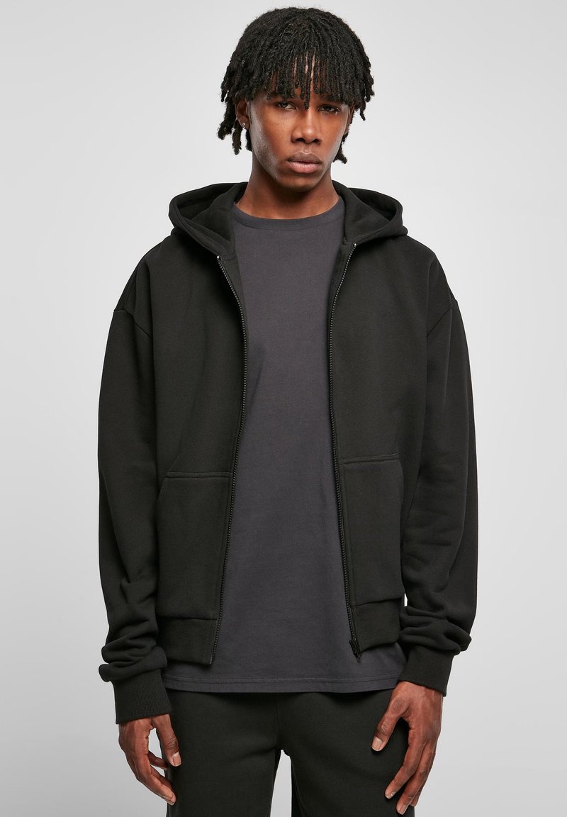 Sweatshirt zippé noir en tissu texturé avec capuche, comportant deux poches avant et des poignets côtelés, superposé à une chemise gris foncé.