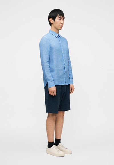 Ανοιχτό μπλε πουκάμισο με μακριά μανίκια και κουμπιά στο μπροστινό μέρος, συνδυασμένο με ναυτικά σορτς. Το μοντέλο φοράει ανοιχτά αθλητικά παπούτσια και μαύρες κάλτσες. Απλός και μοντέρνος σχεδιασμός.
