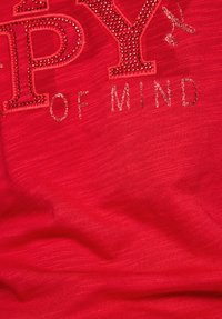 Roter Stoff mit großen "PY"-Buchstaben, verziert mit Strasssteinen und metallischen Akzenten, und dem Satz "OF MIND" in einem helleren metallischen Finish.