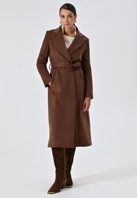 Cappotto in misto lana marrone con cintura, rever a punta e tasche laterali, indossato sopra un maglione beige e abbinato a stivali marroni ricamati al ginocchio.