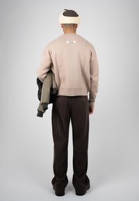 Homme debout de dos, portant un pull beige, un pantalon rayé foncé et un chapeau bordé de fourrure, tenant une veste vert foncé dans une main.