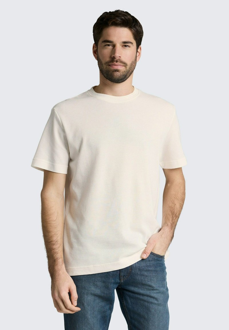 Uomo con capelli scuri e barba, che indossa una maglietta a maniche corte tinta unita color avorio e jeans blu, in piedi con una mano in tasca.