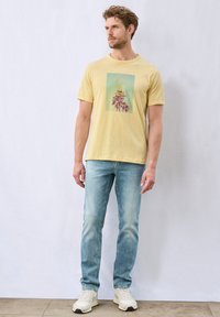 Keltainen puuvillapaita, jossa on graafinen printti palmuista vihreällä taustalla, yhdistettynä vaaleansinisiin farkkuihin ja valkoisiin lenkkareihin.