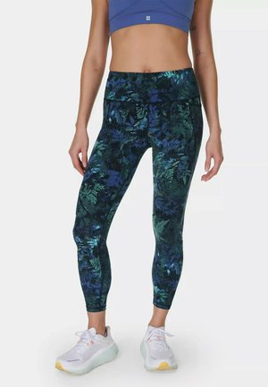 Femme portant un crop top bleu, des leggings à motifs floraux verts foncés et bleus, et des baskets blanches sur un fond clair uni.