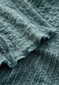 Tekstuuriga kangas vaigistatud teal värvuses, millel on lainelised mustrid ja laineline serv, luues kihilise visuaalse efekti.