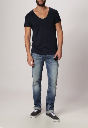 Mørkeblå V-hals t-shirt, blå denim jeans og sorte sneakers. T-shirten har en lomme; jeansene har en falmet vask og let slid.