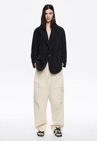 Blazer nero oversize con rever a punta, chiusura a bottone e due tasche frontali, abbinato a pantaloni a gamba larga beige chiaro e sandali a fantasia.