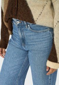 Sinine Denim teksased kõrge vöökohaga, kontrastõmblustega ja klassikalise viie taskuga disain, koos pruunist ja kreemist kootud sviitriga.
