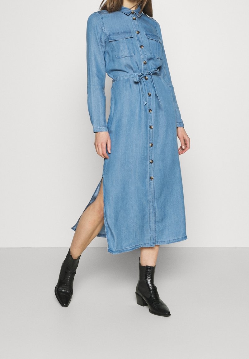 Robe en denim bleu clair avec boutons, à manches longues, deux poches avant, taille ceinturée, fentes latérales et coupe droite. Portée avec des bottines noires.