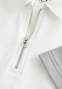 Polo shirt blanc avec zip argenté, col en maille texturée et accents de manches gris. Tissu lisse avec des coutures soignées.