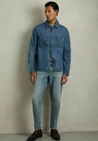 Jachetă din denim de culoare albastru închis, cu două buzunare pe piept, închidere cu nasturi și gulere în vârf, asortată cu blugi de culoare albastru deschis și pantofi negri.