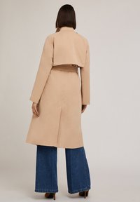 Trench coat beige con dettaglio a mantella sul retro, vita con cintura, maniche lunghe e silhouette fluida, abbinato a pantaloni in denim a gamba larga.