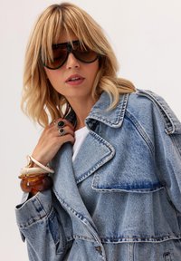 Blondynka w dużych ciemnych okularach przeciwsłonecznych i jasnoniebieskiej kurtce dżinsowej, uzupełniona masywnymi bransoletkami i pierścionkami.