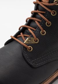 Bota de cuero negro con cordones marrones, ojales dorados y un acabado texturizado. Presenta costuras marrones y un detalle de logo metálico en el costado.