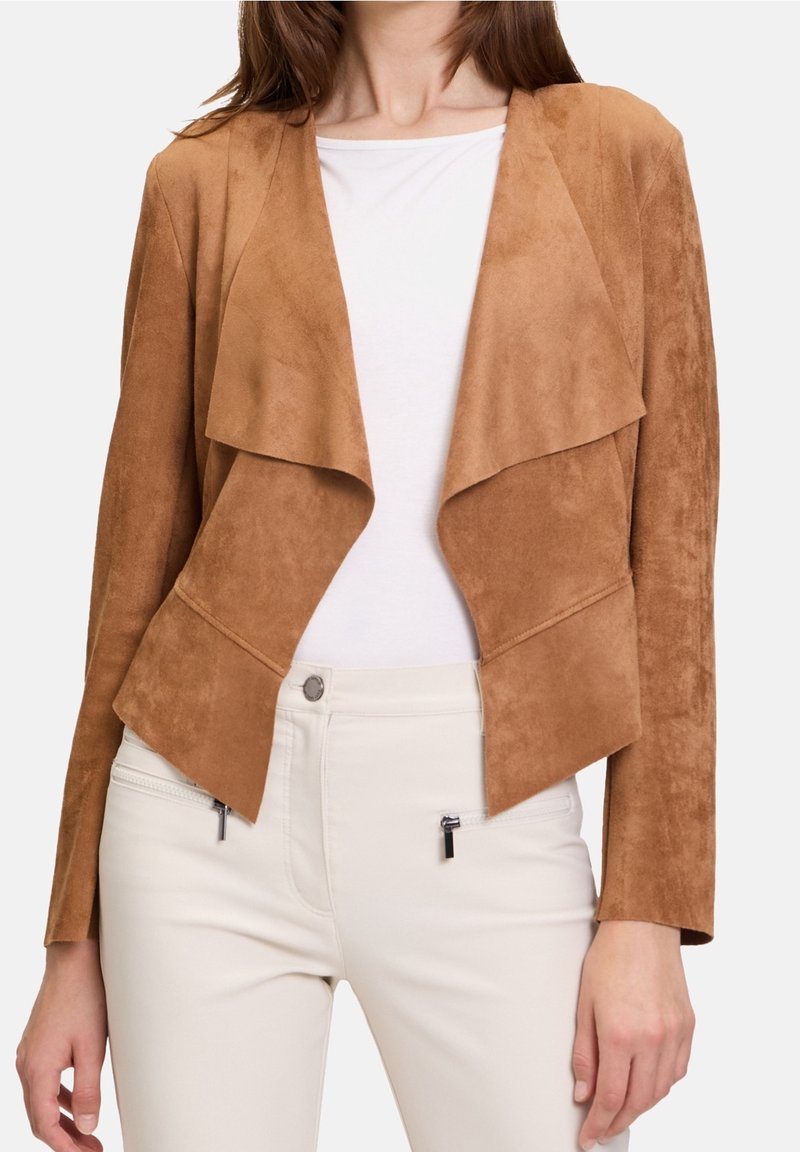 Femme portant une veste en daim beige ouverte sur le devant, par-dessus un haut blanc et un pantalon blanc avec des poches zippées sur un fond clair.