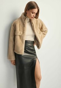 Beżowa kurtka z futra ekologicznego z stójką, kieszeniami na klapie z przodu oraz czarna skórzana spódnica z wysokim rozcięciem, prezentująca gładkie tekstury i kontrastujące kolory.