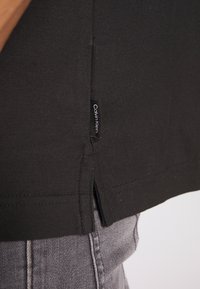 Musta puuvillapaita, jossa on sivuhalkio, ja jossa on pieni Calvin Klein -merkki sauman kohdalla, yhdistettynä harmaisiin farkkuihin.