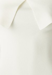 Geripptes Strickoberteil in Off-White mit großem, spitz zulaufendem Kragen. Weiche Textur und figurbetonte Form, die das vertikale Rippendetail hervorhebt.