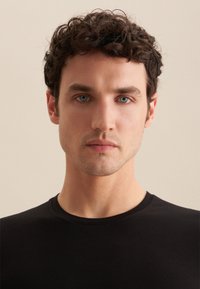 T-shirt noir à col rond en coton. Gros plan sur un mannequin masculin aux cheveux courts et bouclés et aux yeux bleus, sur fond neutre.