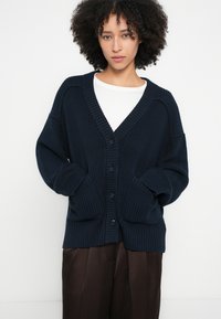 Cardigan bleu marine oversize avec texture côtelée, col en V profond, gros boutons et deux poches avant. Porté avec un haut blanc et un pantalon marron.