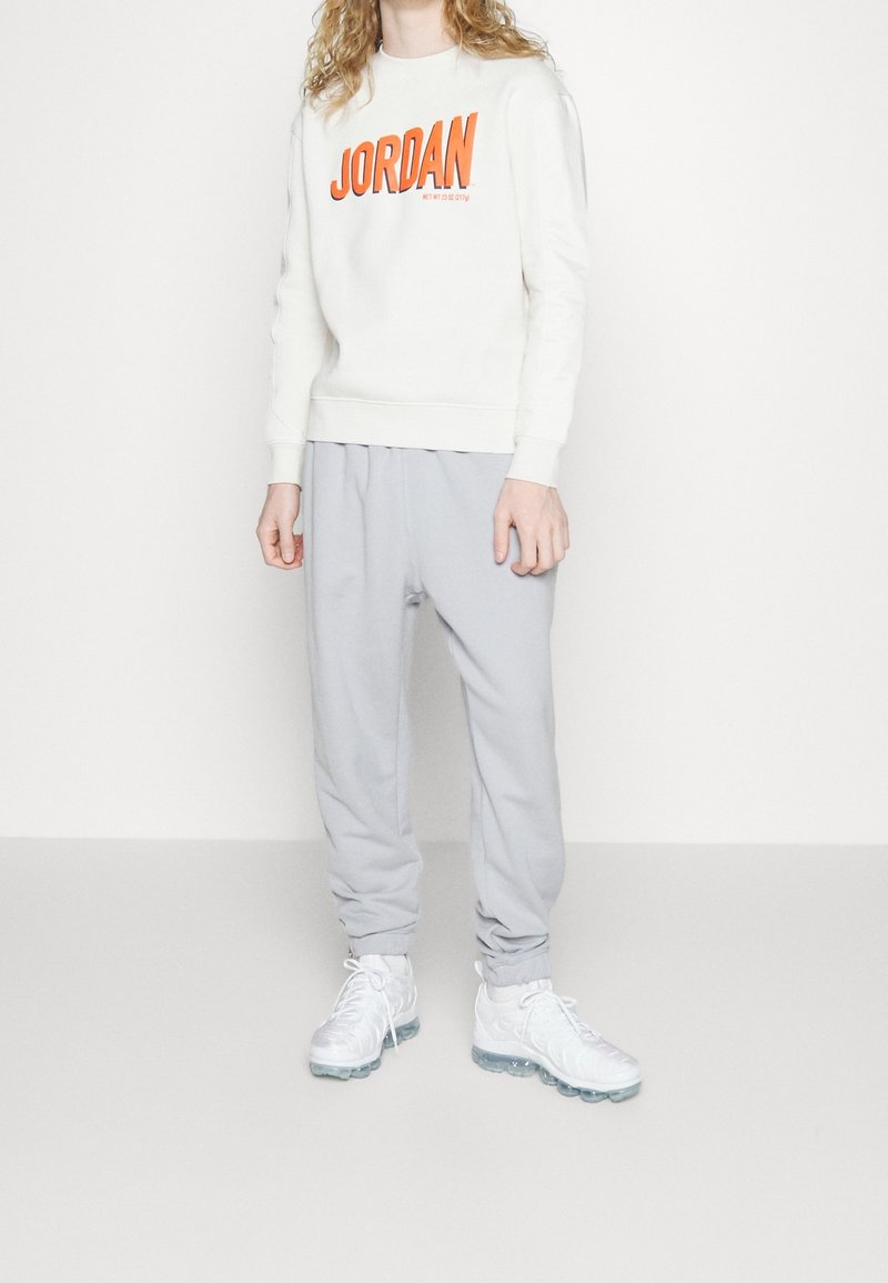 Sweat-shirt blanc avec le texte "JORDAN" en orange, associé à un pantalon de jogging gris clair et des baskets blanches à semelles translucides. Textures de tissu lisses.