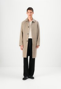 Beige wollen blend overcoat met een kraag, een enkelzijdig ontwerp en zwarte knopen, gedragen over een lichte top en zwarte broek.