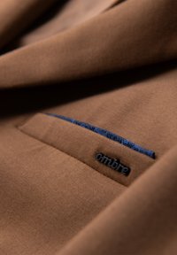Tissu marron avec une poche bordée de bleu et une petite étiquette noire "ombre" sur le bord de la poche.