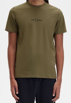Person trägt olivgrünes Fred Perry T-Shirt mit schwarzem Logo auf der Brustmitte und schwarze Hose, steht vor einem einfarbigen Hintergrund.