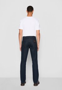 Homme debout de dos, portant un t-shirt blanc à manches courtes, un jean bleu foncé et des chaussures marron, sur un fond clair uni.