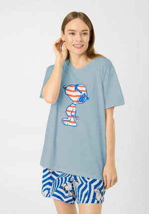 Femme aux longs cheveux portant un T-shirt bleu clair oversize et un short à motifs, tous deux avec des motifs du personnage Snoopy.