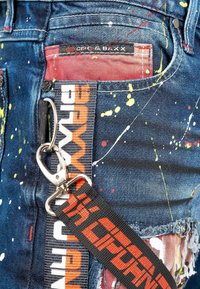 Shorts en denim avec une base bleu foncé ornée de taches de peinture multicolores, des bords effilochés et une sangle tissée noire avec des lettres orange.