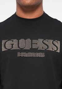 Melnā T-kreklā ar teksturētu beigu "GUESS" logo un detaļu "EST. 1981" uz krūtīm. Apļveida kakla dizains, kokvilnas materiāls.