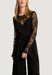 Kobieta ubrana w czarną koronkową bluzkę z długim rękawem i kwiatowymi wzorami, w połączeniu z czarnymi szerokimi spodniami z wysokim stanem, stojąca na tle o neutralnym kolorze.