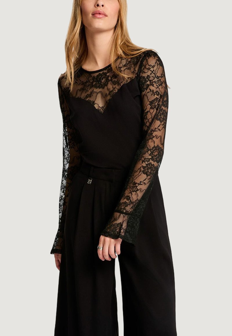 Kobieta ubrana w czarną koronkową bluzkę z długim rękawem i kwiatowymi wzorami, w połączeniu z czarnymi szerokimi spodniami z wysokim stanem, stojąca na tle o neutralnym kolorze.