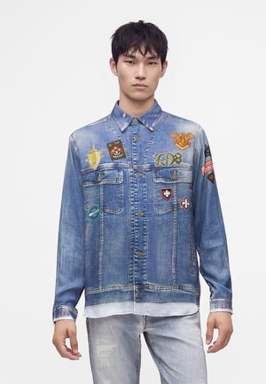 Jeune homme portant une veste en jean bleue avec plusieurs patches colorés et un jean gris clair, debout devant un fond blanc uni.