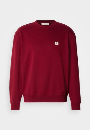 Punane sviiter, mis on valmistatud pehmest kangast, millel on ümmargune kaelus, pikad varrukad ja väike brodeeritud logo vasakul rinnakorvil.