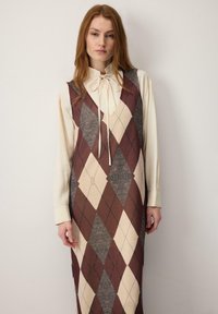 Dolga argyle obleka z vzorci rjavih, kremnih in sivih diamantov, preko lahke bež bluze z zavezanim izrezom na vratu.