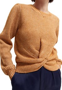 Pull en tricot orange doré, avec un design torsadé à l'avant, manches longues et texture côtelée. Associé à un pantalon bleu foncé.