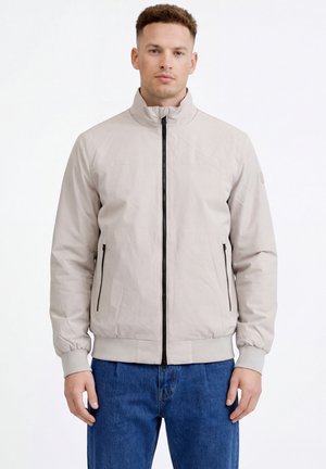 Uomo che indossa una giacca beige chiaro con cerniera e collo alto, e jeans blu, in piedi davanti a uno sfondo bianco uniforme.