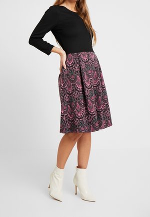 Vrouw die een zwart topje met lange mouwen draagt, een knielange, roze-zwart patroonrok en witte enkellaarzen met hakken, staand met één hand op de heup.